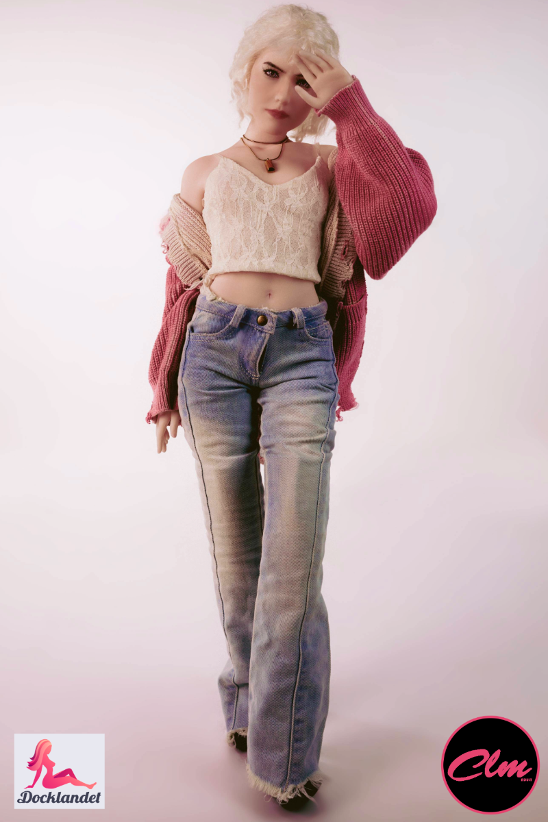 sexdukke Tara realistisk mini 60 cm i silikon, blond dukke i jeans, crop top og rosa jakke, stående oppreist mot lys studiobakgrunn.